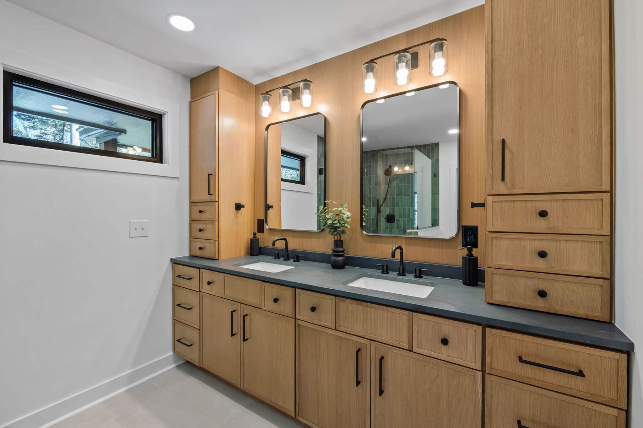 Remodeled bathroom with a white oak double vanity, dark stone counter and a green tiled shower